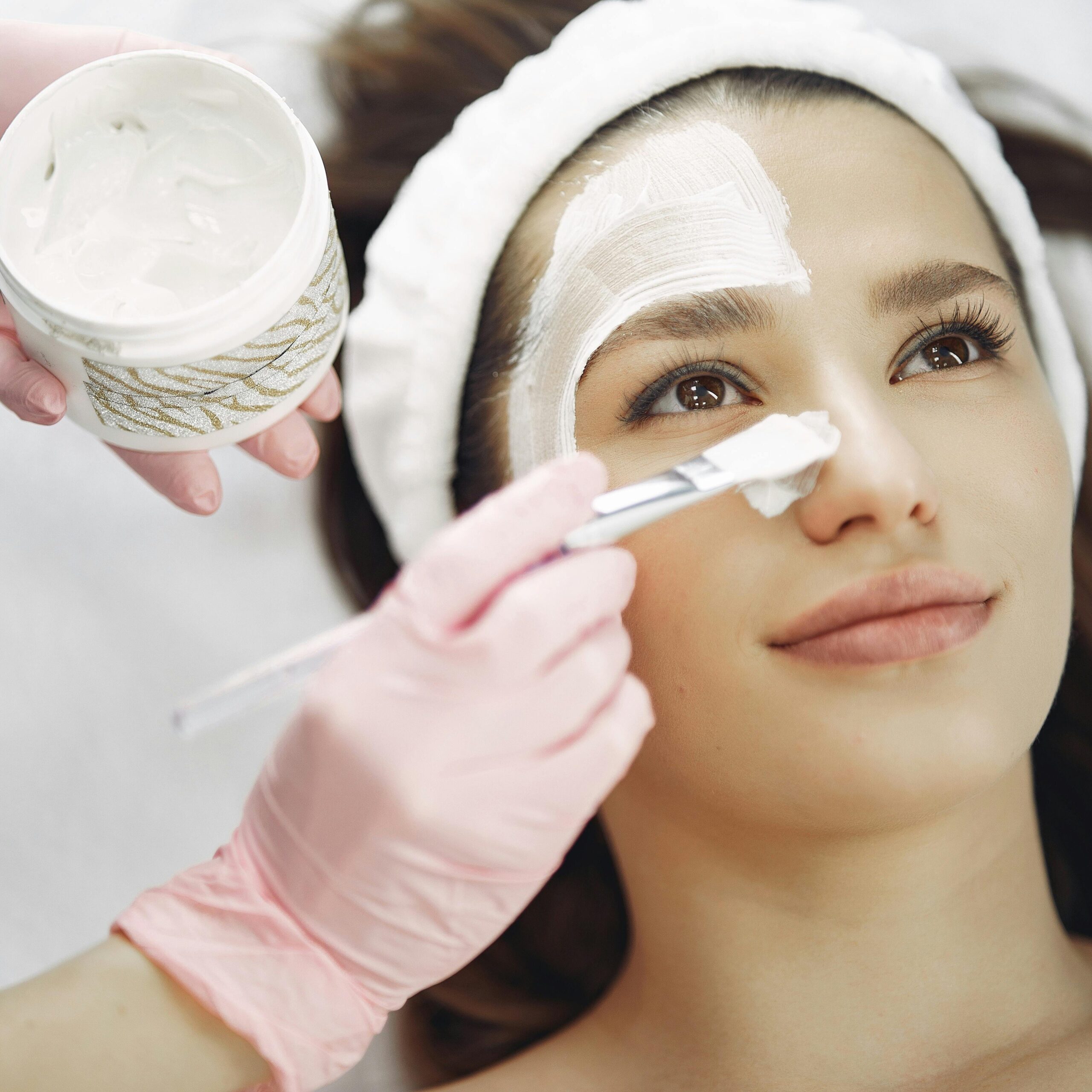 Close-up of a woman receiving a facial treatment at a spa with a cream mask applied using a brush.